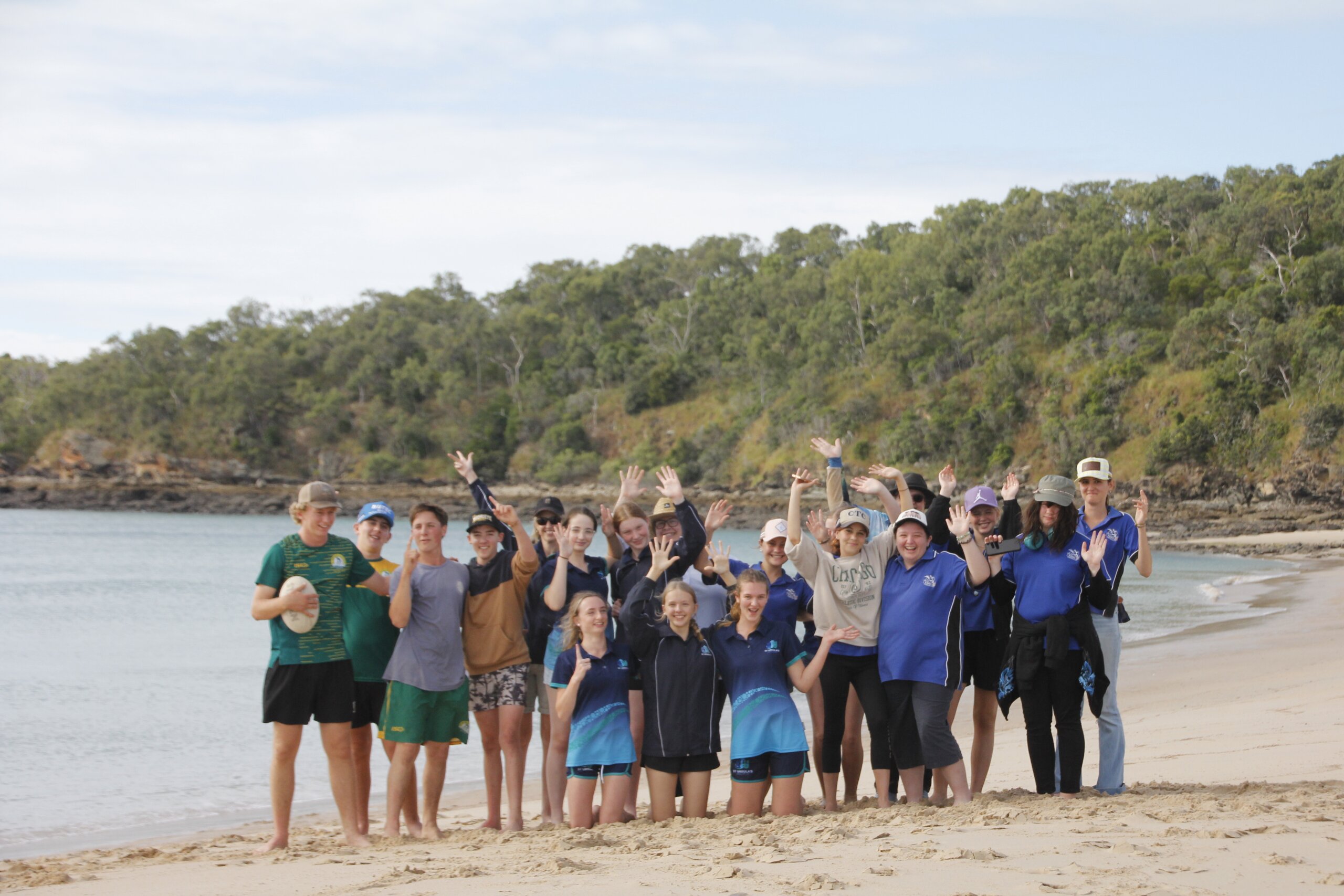 A group of teenagers standing on the beach waving at the camera